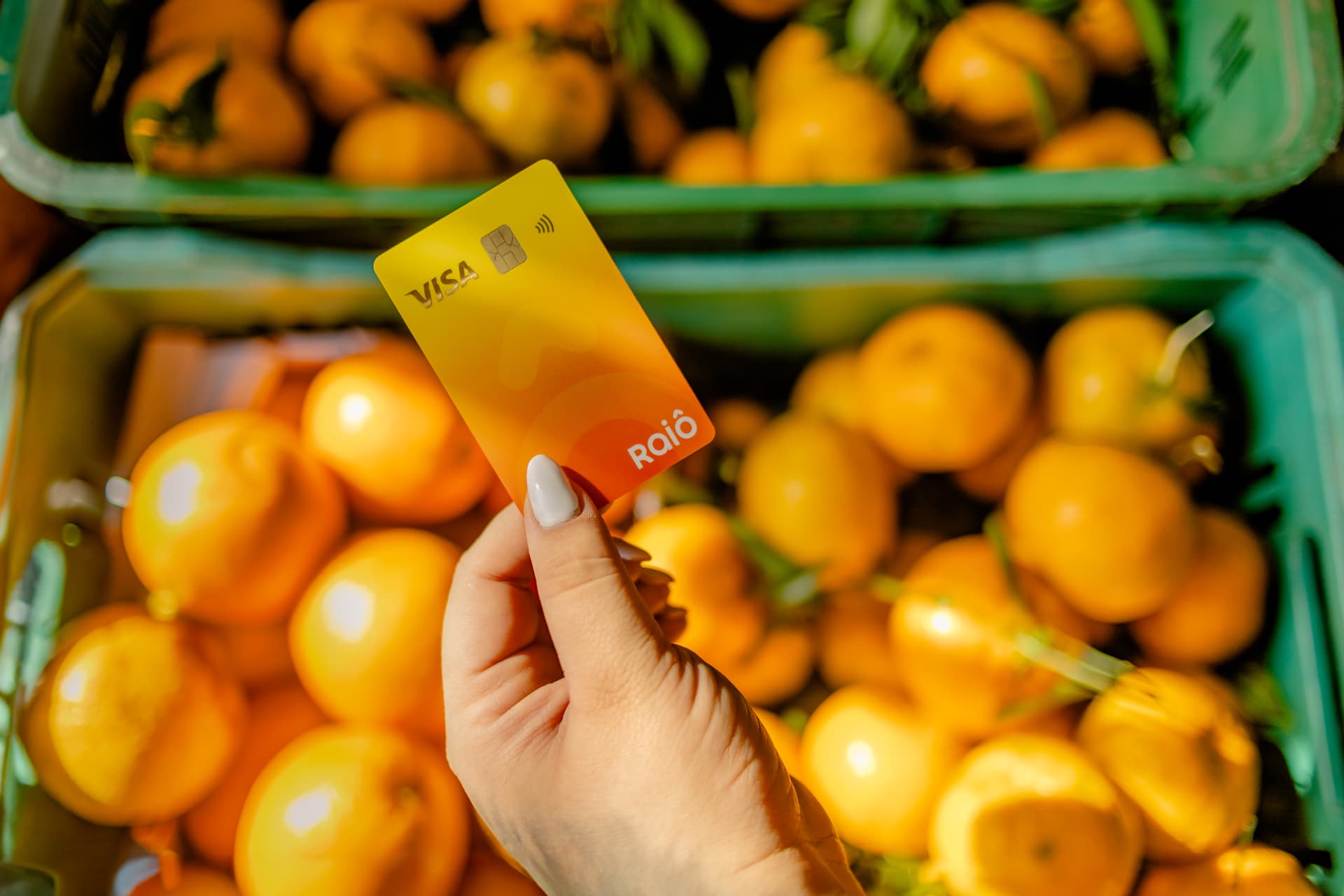 Mão segurando um cartão Raiô Visa em frente a caixas de laranja colhidas
